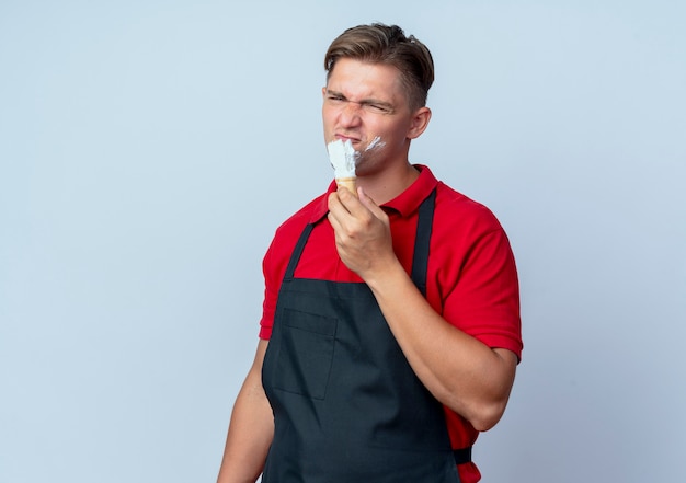 Young annoyed blonde male barber in uniform smears shaving foam on face isolated on white space with copy space