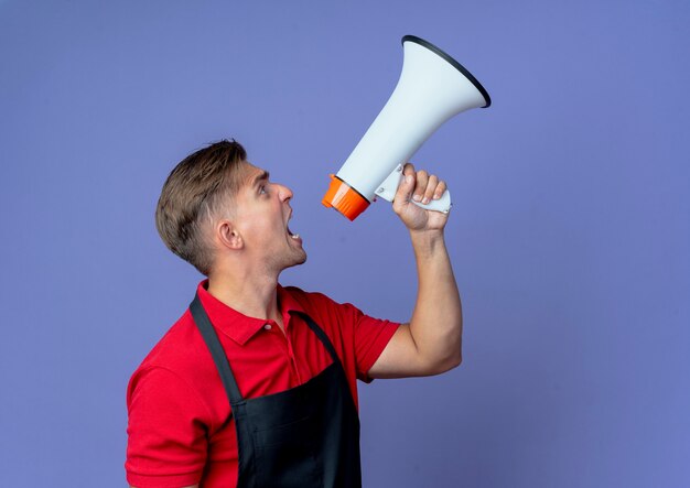 Young annoyed blonde male barber in uniform stands sideways shouts into loud speaker looking up isolated on violet space with copy space