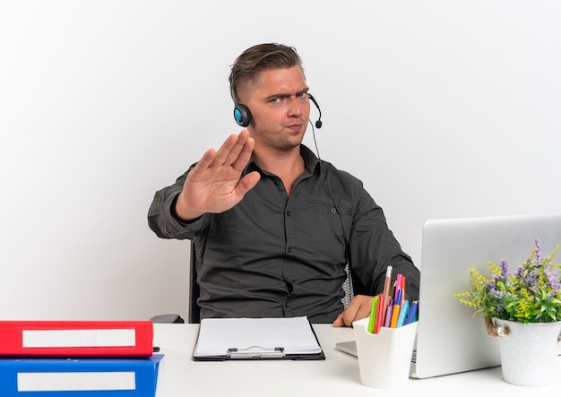 Young annoyed blonde office worker man on headphones sits at desk with office tools using laptop gestures stop hand sign isolated on white background with copy space