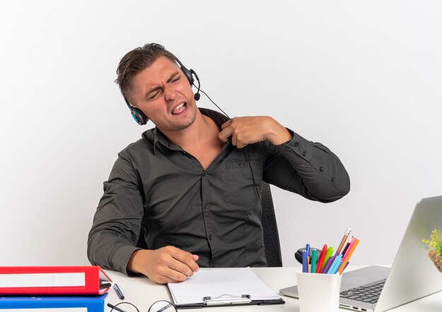 Young annoyed blonde office worker man on headphones sits at desk with office tools using laptop holds collar isolated on white background with copy space