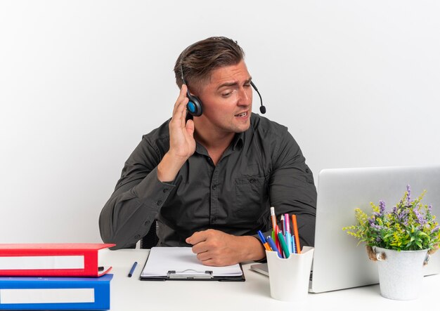 Young annoyed blonde office worker man on headphones sits at desk with office tools using laptop holds headphones trying to hear isolated on white background with copy space