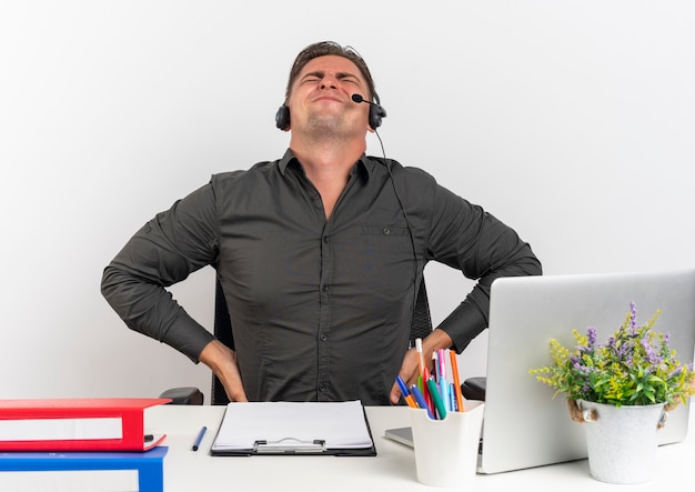 Foto gratuita giovane uomo di lavoratore di ufficio bionda infastidito sulle cuffie si siede alla scrivania con strumenti per ufficio utilizzando il computer portatile tiene la schiena guardando in alto isolato su sfondo bianco con spazio di copia