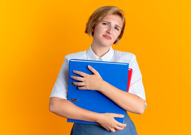 La giovane ragazza russa bionda infastidita tiene le cartelle di file isolate su priorità bassa arancione con lo spazio della copia