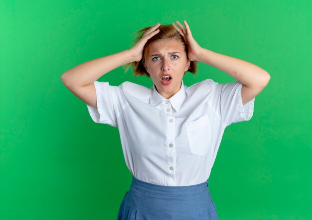 Young annoyed blonde russian girl holds head isolated on green background with copy space