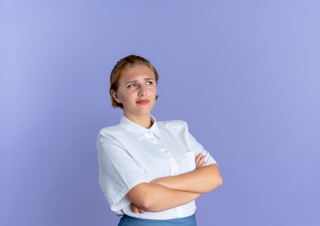 Молодая раздраженная русская блондинка смотрит в сторону со скрещенными руками, изолированными на фиолетовом фоне с копией пространства