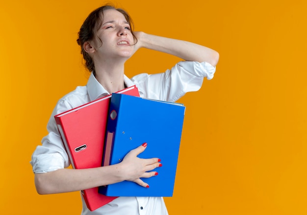 Young annoyed blonde russian girl puts hand on head behind holding file folders on orange  with copy space