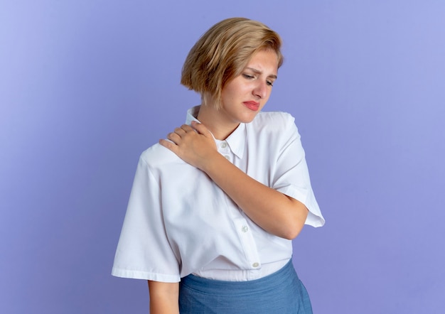 Free photo young annoyed blonde russian girl puts hand on shoulder looking down isolated on purple background with copy space