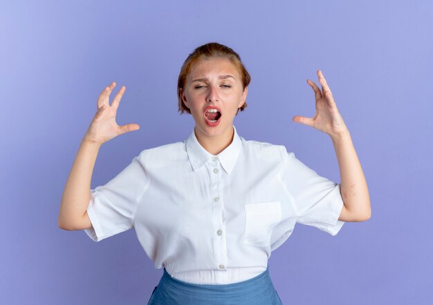 Young annoyed blonde russian girl stands with raised hands isolated on purple background with copy space