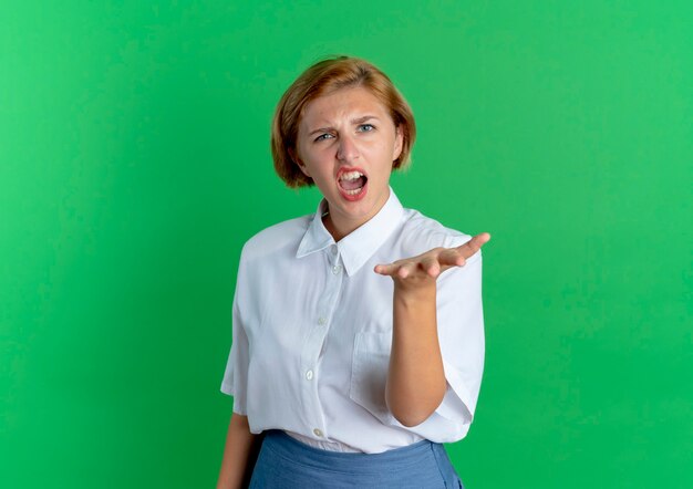 Young annoyed blonde russian girl yells and points at camera isolated on green background with copy space