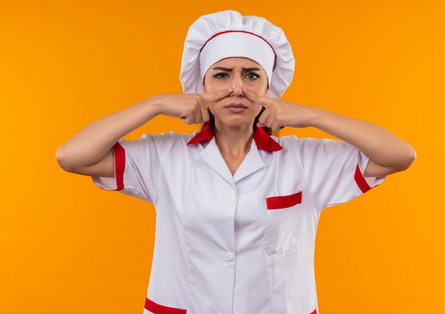 Молодая раздраженная кавказская девушка-повар в униформе шеф-повара закрывает нос пальцами, изолированными на оранжевом фоне с копией пространства