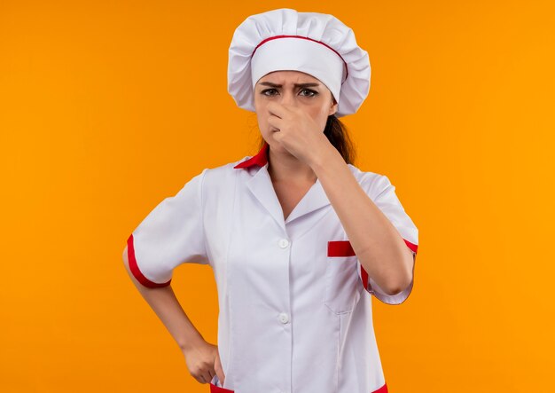 Молодая раздраженная кавказская девушка-повар в униформе шеф-повара закрывает нос рукой, изолированной на оранжевом фоне с копией пространства