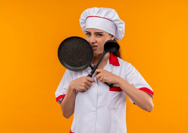 Молодая раздраженная кавказская девушка-повар в униформе шеф-повара пересекает сковороду и ковш, изолированные на оранжевом фоне с копией пространства