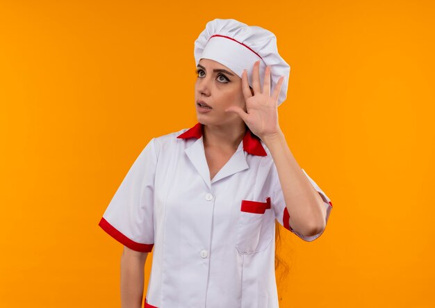 Young annoyed caucasian cook girl in chef uniform gestures can't hear sign isolated on orange background with copy space