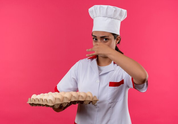 Young annoyed caucasian cook girl in chef uniform holds batch of eggs and closes nose isolated on pink background with copy space