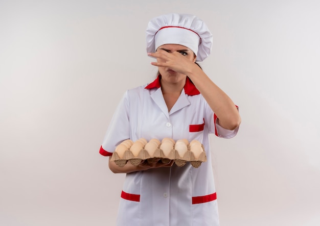 Foto gratuita la giovane ragazza caucasica infastidita del cuoco in uniforme del cuoco unico tiene il lotto delle uova e chiude il naso con la mano isolata su fondo bianco con lo spazio della copia