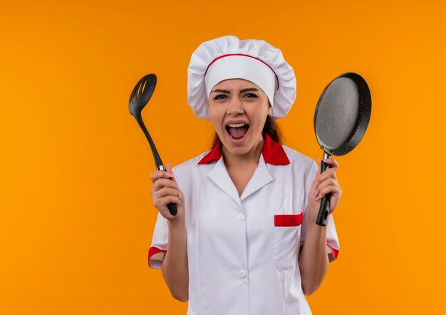 Молодая раздраженная кавказская девушка-повар в униформе шеф-повара держит сковороду и лопатку, изолированные на оранжевом фоне с копией пространства