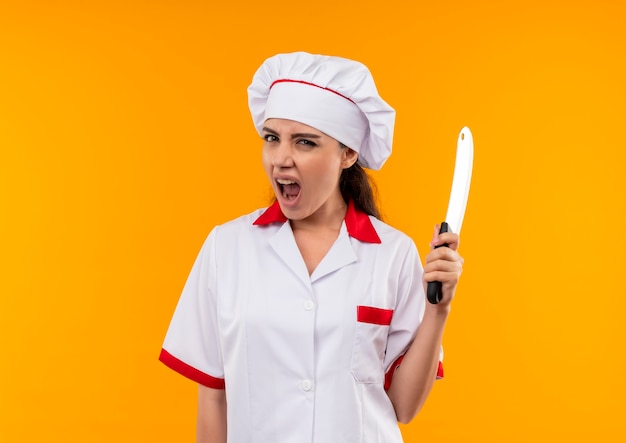 La giovane ragazza caucasica infastidita del cuoco in uniforme del cuoco unico tiene il coltello isolato su priorità bassa arancione con lo spazio della copia