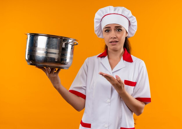 Young annoyed caucasian cook girl in chef uniform holds and points with hand at pot isolated on orange background with copy space