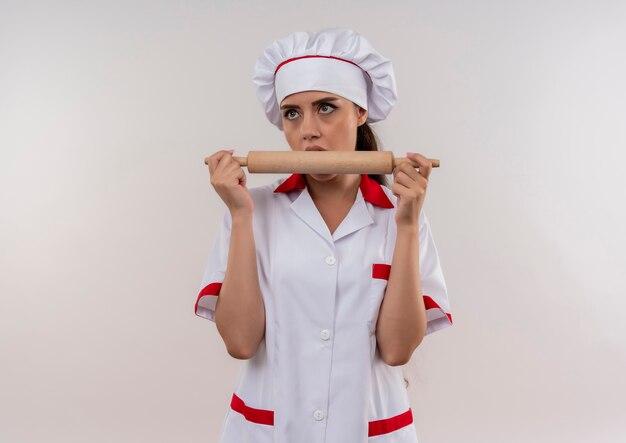 Young annoyed caucasian cook girl in chef uniform holds and pretends to bite rolling pin isolated on white background with copy space