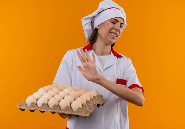 Young annoyed caucasian cook girl in chef uniform holds and pretends to push with hand batch of eggs isolated on orange background with copy space