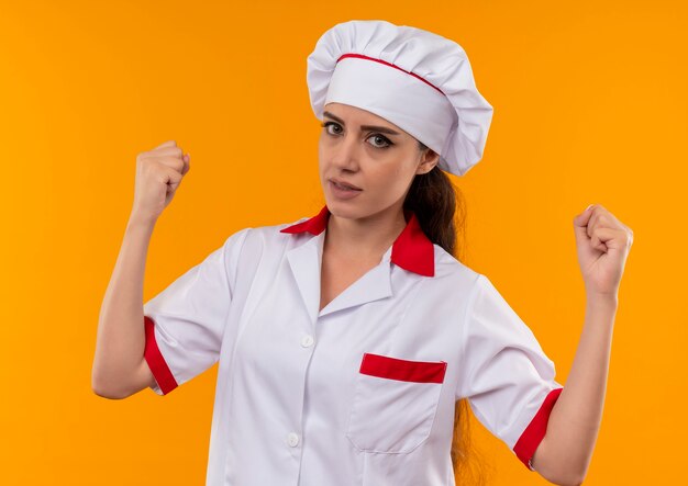 Young annoyed caucasian cook girl in chef uniform raises fists up isolated on orange background with copy space