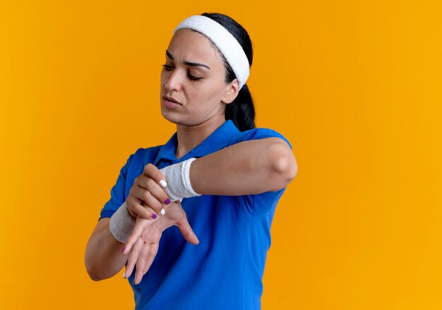 Young annoyed caucasian sporty woman wearing headband and wristbands holds hand on orange  with copy space