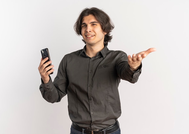 Молодой раздраженный красивый кавказский мужчина держит телефон и указывает на камеру, изолированную на белом фоне с копией пространства