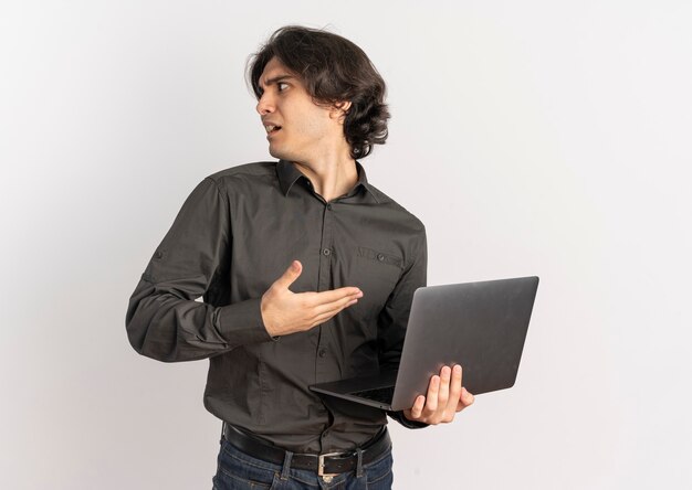 Young annoyed handsome caucasian man holds and points at laptop looking at side isolated on white background with copy space