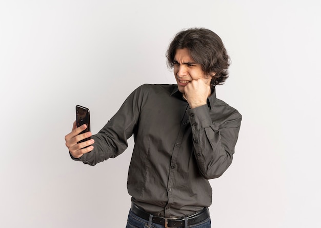Young annoyed handsome caucasian man puts fist on face and looks at phone isolated on white background with copy space