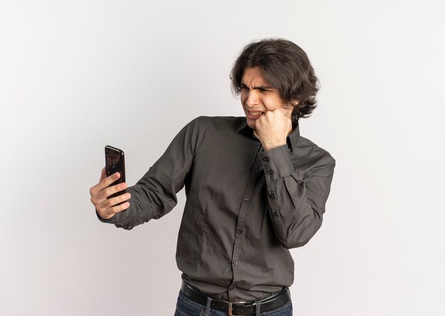 Молодой раздраженный красивый кавказский мужчина кладет кулак на лицо и смотрит на телефон, изолированный на белом фоне с копией пространства