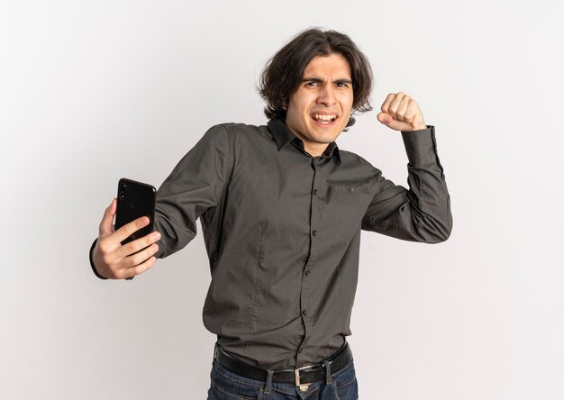 Young annoyed handsome caucasian man raises fist up and holds phone looking at camera isolated on white background with copy space