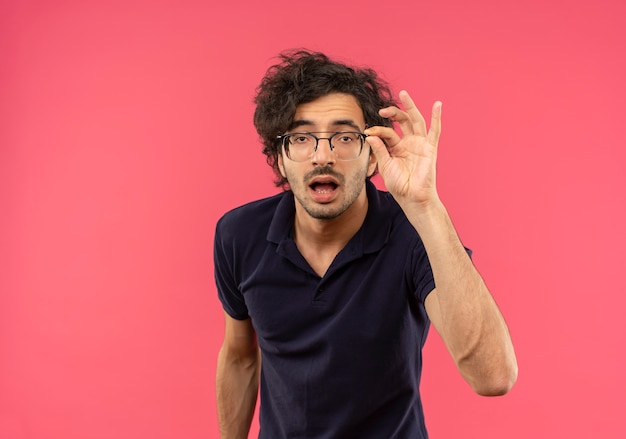 Free photo young annoyed man in black shirt with optical glasses holds glasses and looks isolated on pink wall