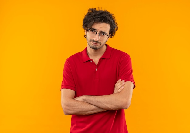 Young annoyed man in red shirt with optical glasses crosses arms and looks isolated on orange wall