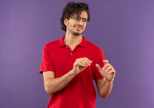 Молодой раздраженный мужчина в красной рубашке с оптическими очками держит руки вверх изолированными на фиолетовой стене