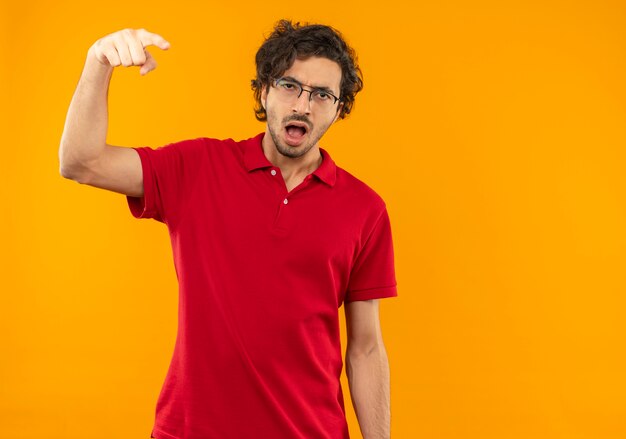 Young annoyed man in red shirt with optical glasses points and looks isolated on orange wall