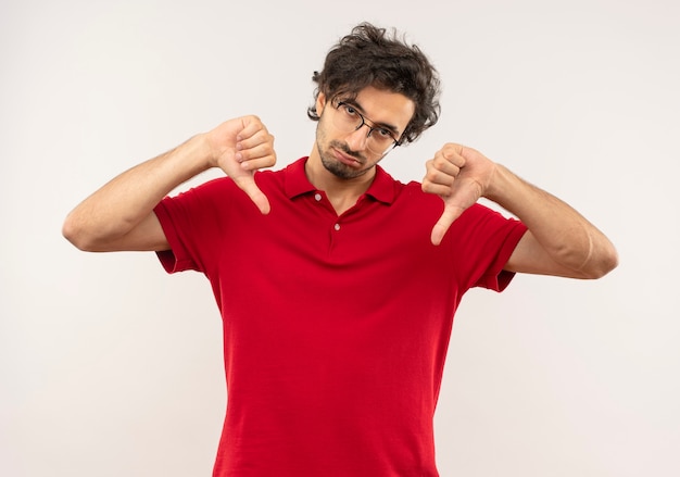 Молодой раздраженный мужчина в красной рубашке с оптическими очками опускает палец вниз двумя руками, изолированными на белой стене