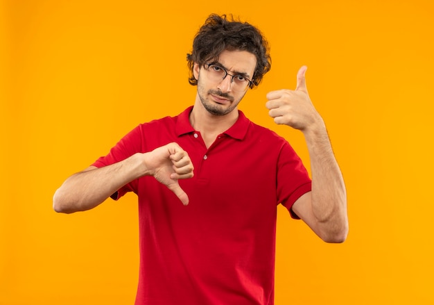 Young annoyed man in red shirt with optical glasses thumbs up and thumbs down isolated on orange wall