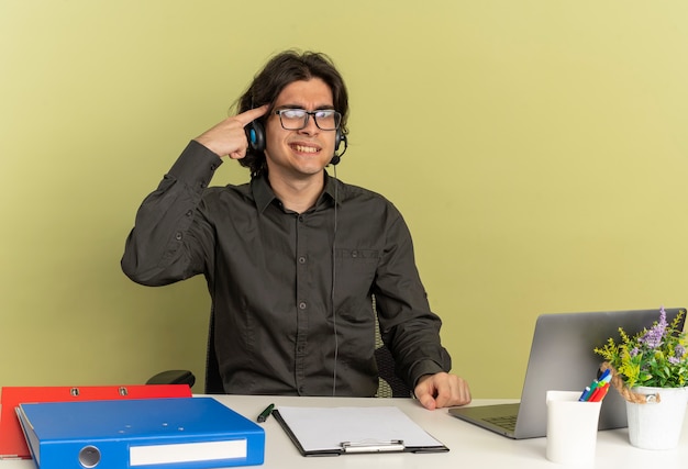 Молодой раздраженный человек офисного работника в наушниках в оптических очках сидит за столом с офисными инструментами, используя ноутбук, кладет палец на голову, глядя в камеру, изолированную на зеленом фоне с копией пространства