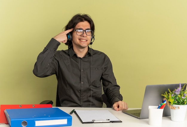 Young annoyed office worker man on headphones in optical glasses sits at desk with office tools using laptop puts finger on head looking at camera isolated on green background with copy space