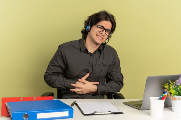 Молодой раздраженный человек офисного работника в наушниках в оптических очках сидит за столом с офисными инструментами, используя ноутбук, кладет руки на живот, изолированные на зеленом фоне с копией пространства