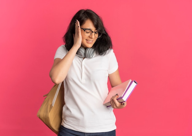 Young annoyed pretty caucasian schoolgirl wearing glasses back bag and headphones on neck closes ear with hand holding book and notebook on pink  with copy space