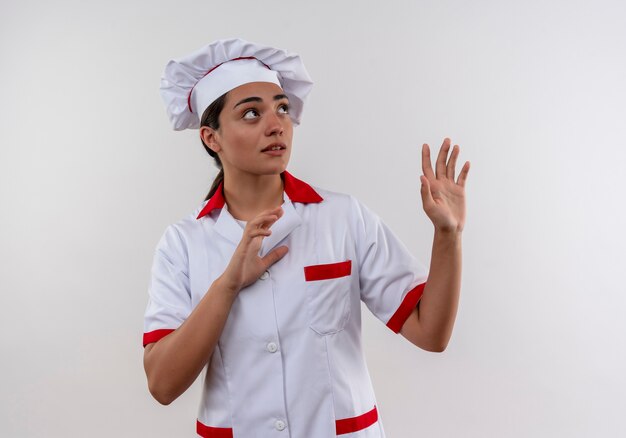 Young anxiois caucasian cook girl in chef uniform pretends to defend with hands and looks up isolated on white background with copy space