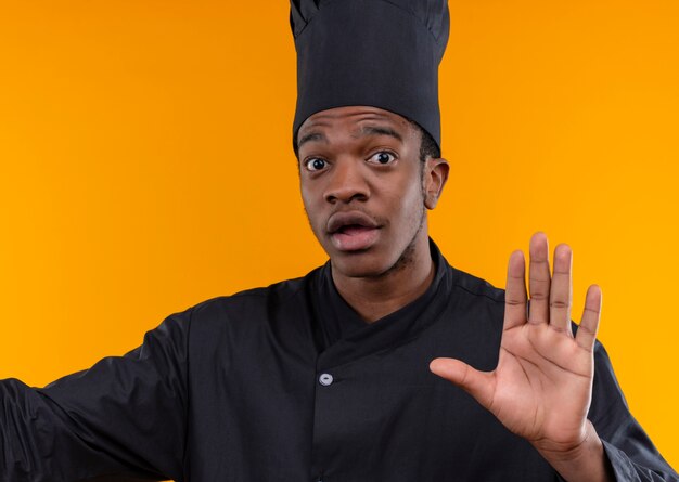 シェフの制服のジェスチャーで若い気になるアフリカ系アメリカ人の料理人は、コピースペースでオレンジ色の背景に分離された手のサインを停止します