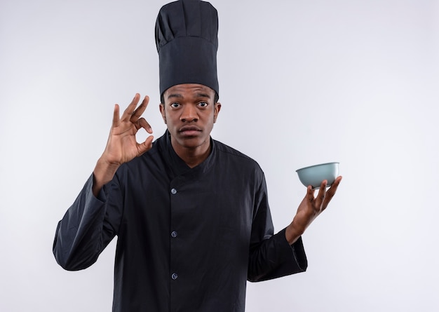 Il giovane cuoco afroamericano ansioso in uniforme del cuoco unico tiene il segno giusto della mano di gesti e della ciotola isolato su fondo bianco con lo spazio della copia