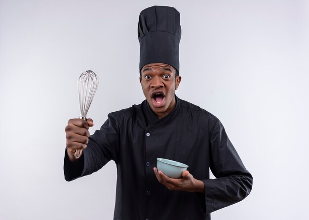 Young anxious afro-american cook in chef uniform holds bowl and whisk isolated on white background with copy space