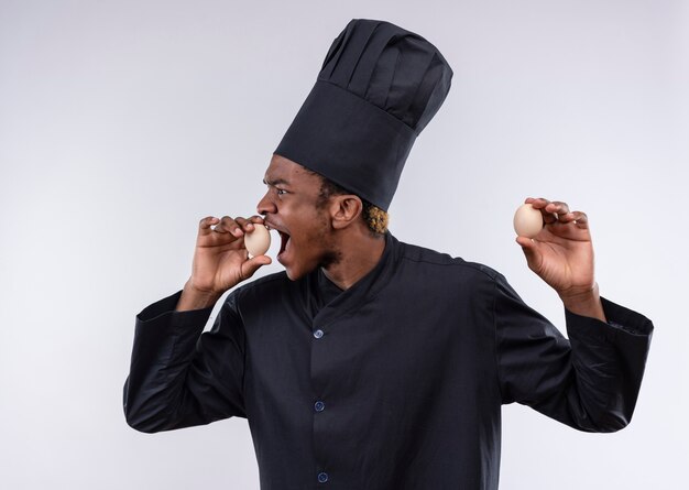 Молодой взволнованный афро-американский повар в униформе шеф-повара держит яйца обеими руками, изолированными на белом фоне с копией пространства