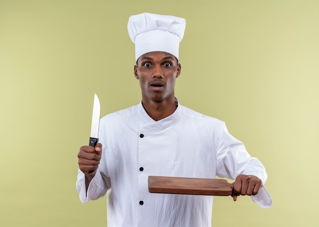 Молодой взволнованный афро-американский повар в униформе шеф-повара держит нож и деревянный кухонный стол, изолированные на зеленом фоне с копией пространства