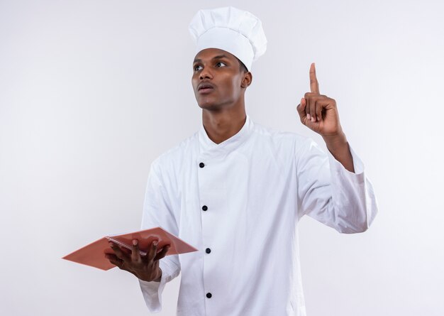 Young anxious afro-american cook in chef uniform holds notebook and points up isolated on white background with copy space