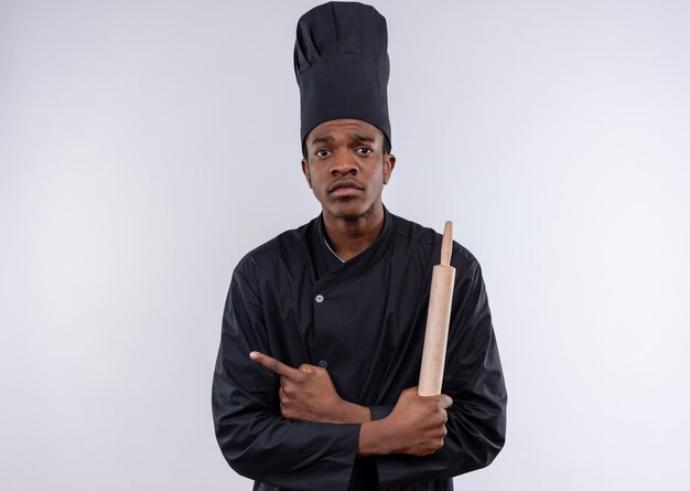 Young anxious afro-american cook in chef uniform holds rolling pin and points to the side isolated on white background with copy space