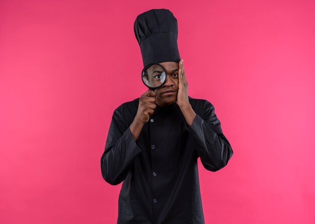 Young anxious afro-american cook in chef uniform looks through magnifying glass or loupe isolated on pink background with copy space